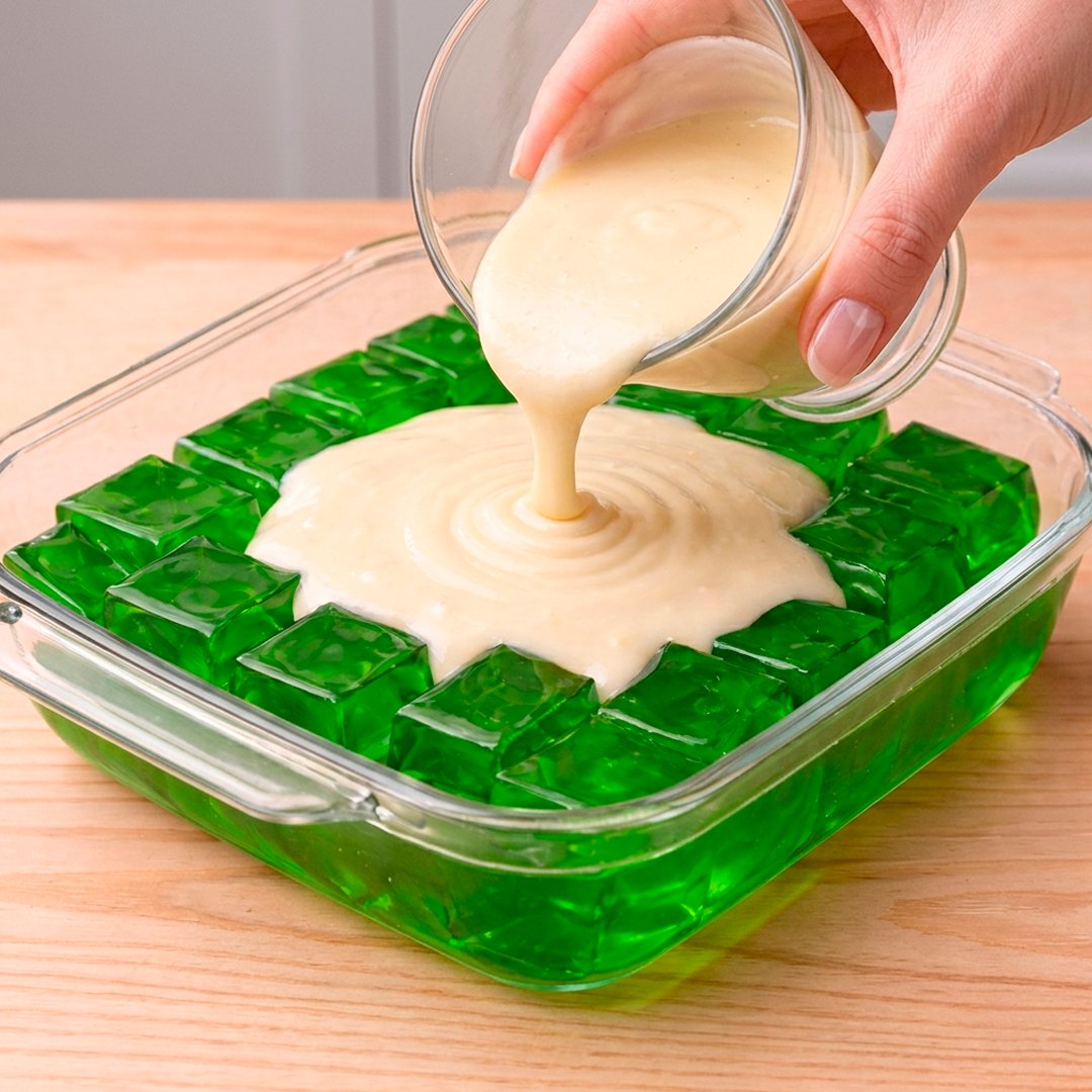 Hand pouring condensed milk vanilla cream over vivid green jelly cubes in a glass dish